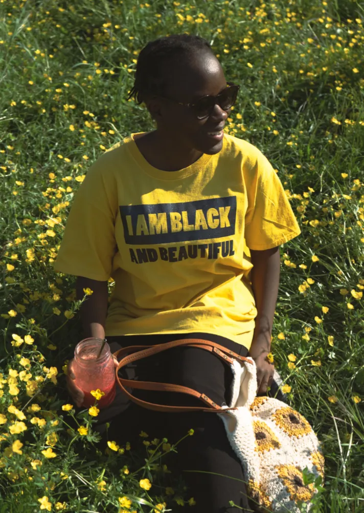Femme dans les champs portant un t-shirt jaune avec le texte “I am Black and Beautiful” à l'avant