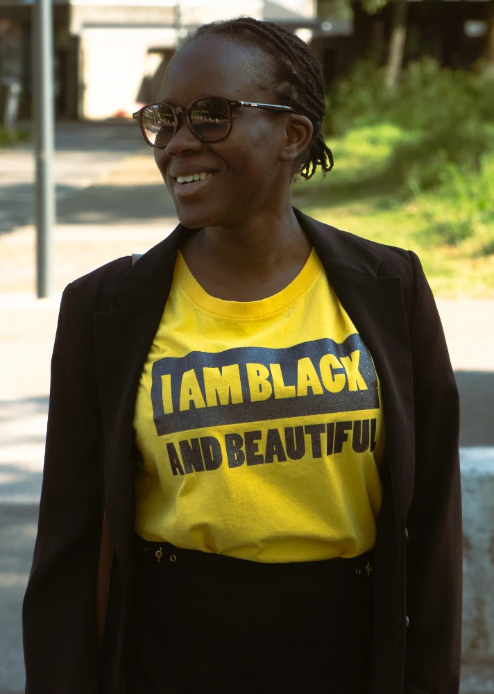 Femme dans la rue portant un t-shirt jaune avec le texte “I am Black and Beautiful” à l'avant